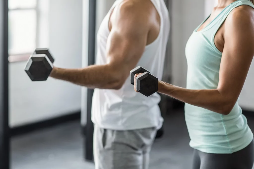 Man and woman performing upper-body dumbbell exercises that can prevent breast sagging, focusing on arm and chest muscle strengthening in a gym setting.