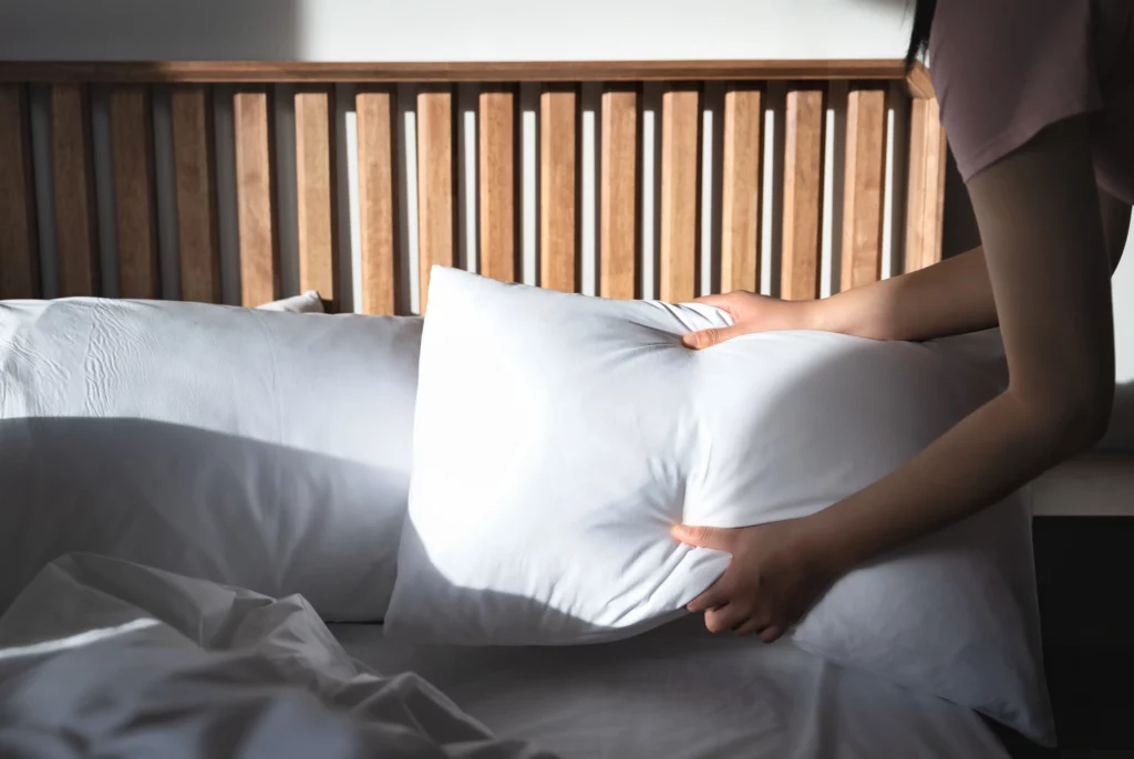 Woman fluffing a white pillow on a bed — arranging extra pillows is a key step in breast augmentation recovery prep.