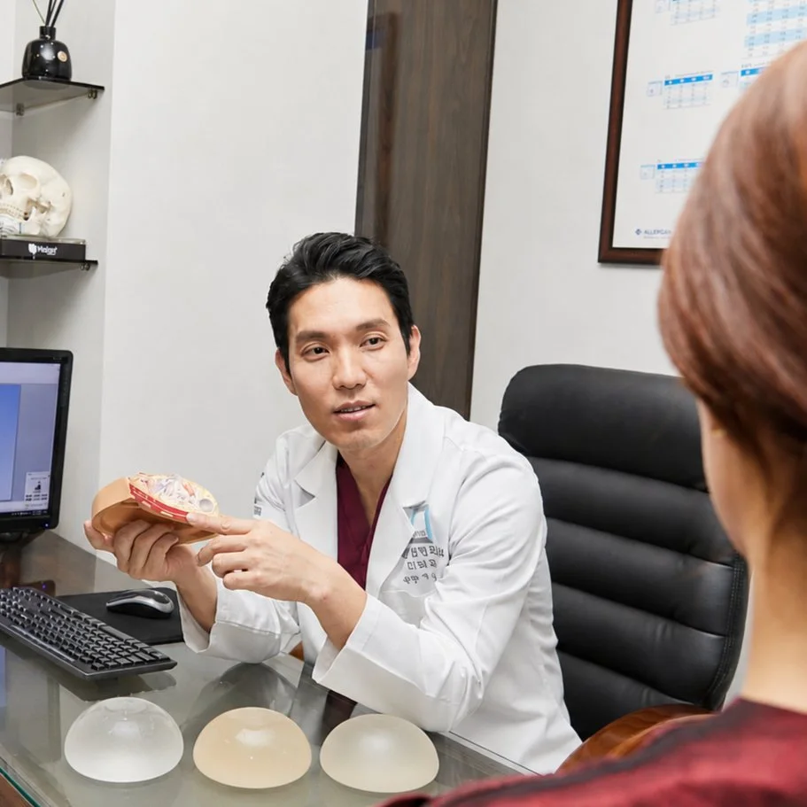 Surgeon holding a breast model with silicone implant samples on the table during a breast implant exchange consultation.