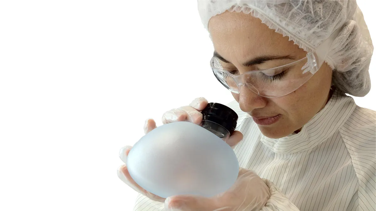 A technician in a cleanroom inspects a silicone breast implant with a magnifying loupe during manufacturer quality control.